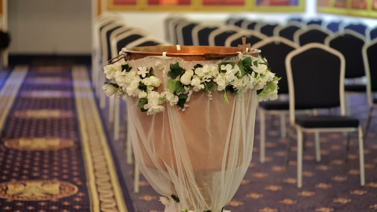 Left-to-right slider shot showing decorated baptism font, cross blur in foreground.