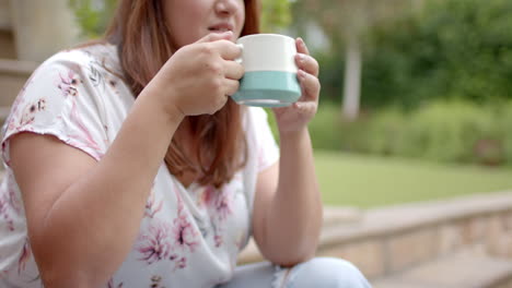 feliz tamaño más mujer biracial sentada en los escalones en el jardín bebiendo café, espacio de copia, cámara lenta