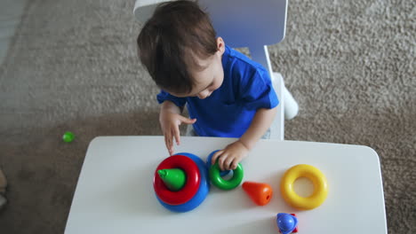 dark-haired caucasian child assembling toy pyramid. charming kid plays sitting at desk indoors. top view.