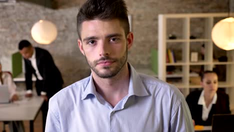 young calm businessman is watching at camera in office, his colleagues are networking with technologies, working concept, business concept, communication concept, blurred background