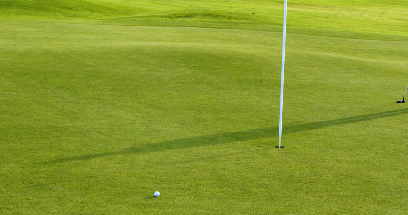 Un'asta della bandiera si trova nella buca su un green di un campo da golf ben curato, proiettando una lunga ombra sull'erba verde vibrante in una giornata di sole.