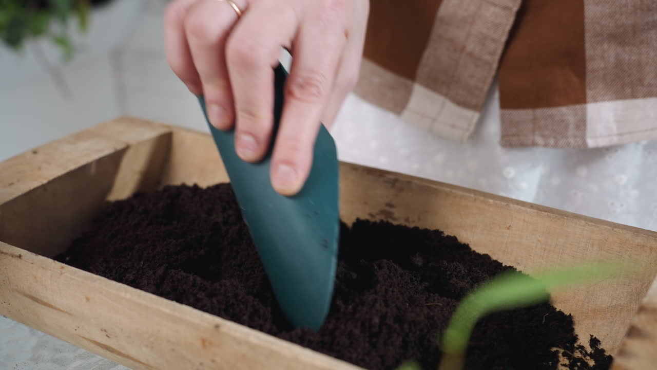 Hand view of farmer using scoop to mix rich dark soil in wooden planter bucket on glass table surrounded by potted plant ceramic tools and natural light in cozy indoor garden space