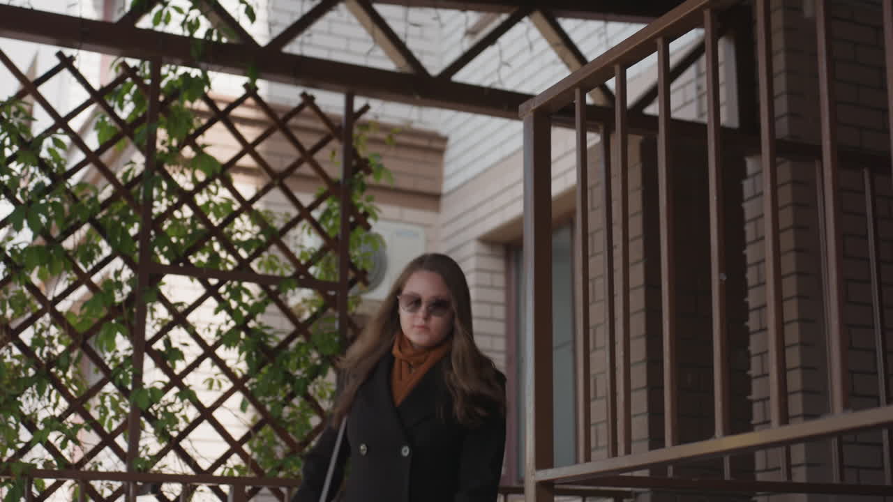 어두운 코트와 안경을 쓴 젊은 여성이 벽돌 난간과 신선한 아이비가 있는 나무 격자 옆 외부 계단을 자신감 넘치는 걸음으로 내려오고 있어요. 도시적인 현관 풍경, 부드러운 햇빛, 차분한 분위기, 은은한 깊이감이 느껴지네요.