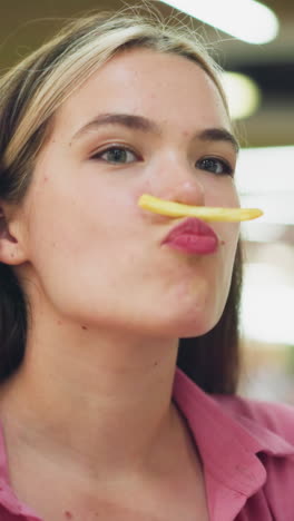 dama blanca en vestido rosa sentada en un restaurante coloca juguetón chip de patata en su labio superior como un bigote, luego lo quita mientras sonríe, con luz borrosa suave y personas en el fondo