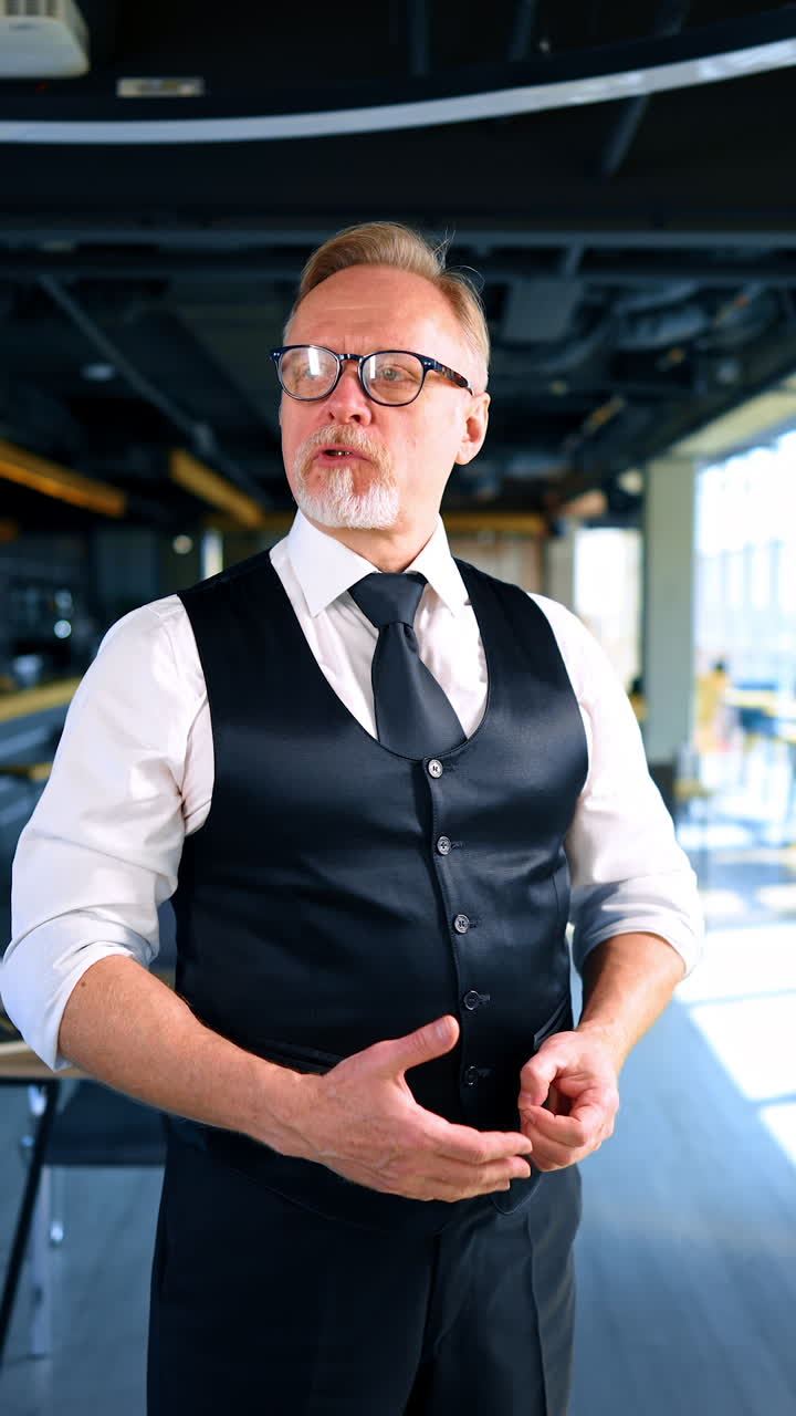 Elegant senior male in white shirt, black vest and trousers in office. Man talks looking around. Vertical video.