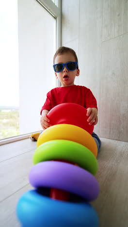 small caucasian boy sitting on the floor puts on sunglasses. toddler takes a toy pyramid and disassembles it. vertical video.