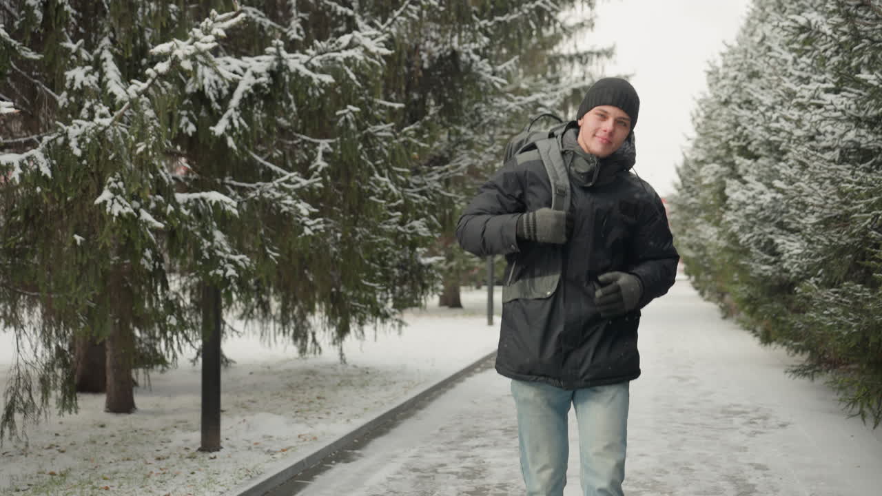 Caucasian man walking with backpack on snowy path, purposeful stride through evergreen avenue, winter jacket and beanie, urban commute vibe with crisp cold light and determined expression