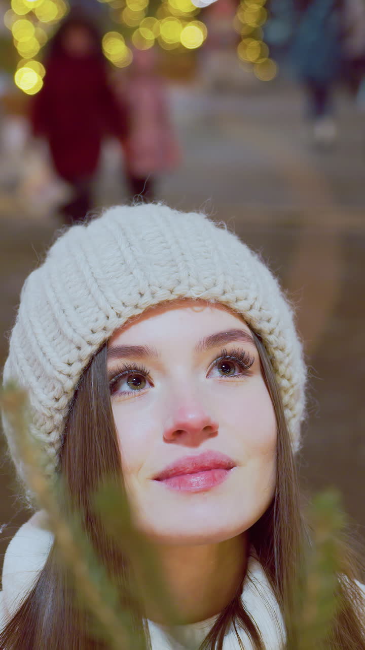 Smiling woman in white beanie and winter coat admiring pine tree with warm holiday glow, soft festive lights highlight her face while people pass by in blurred background