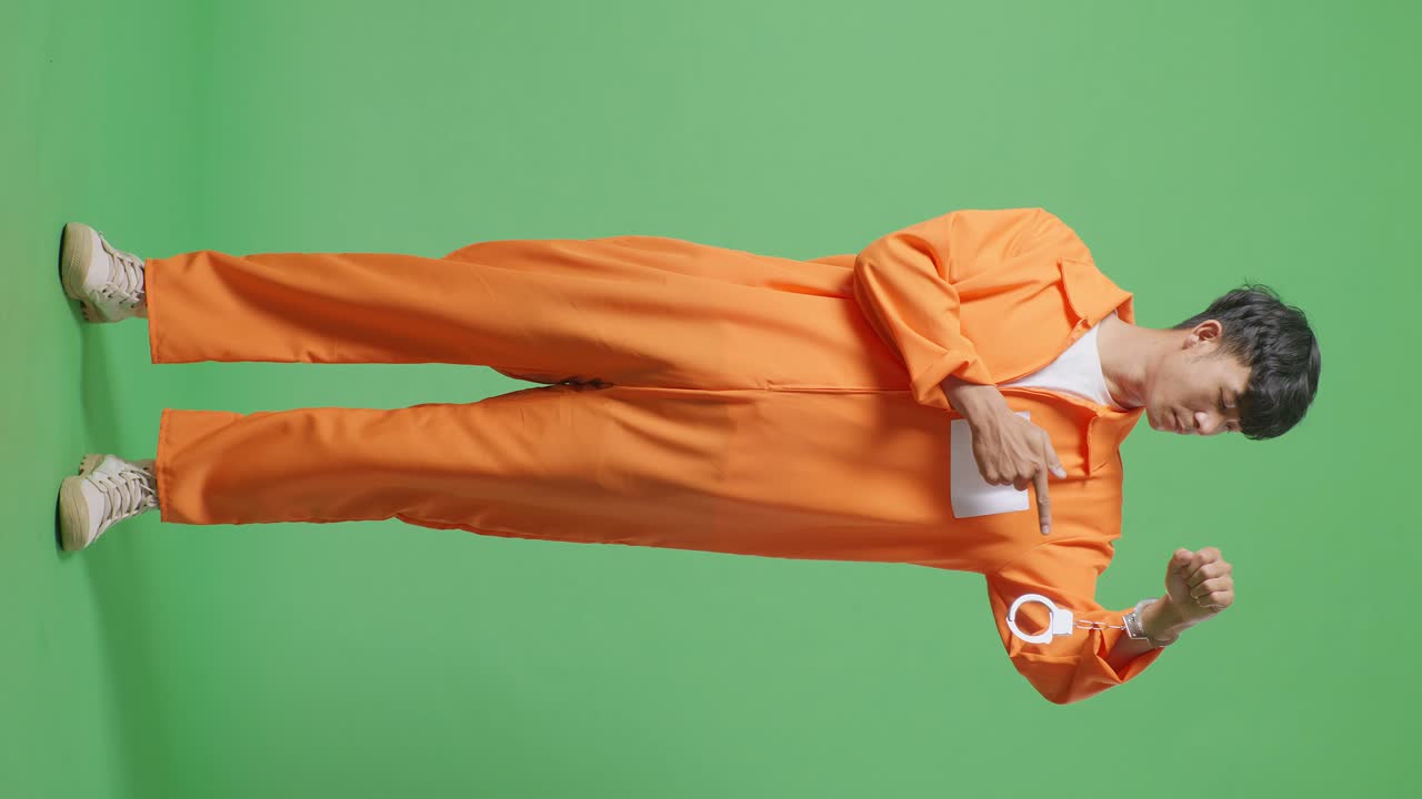 Full Body Of Asian Male Prisoner Standing On The Green Screen Background, Showing The Handcuffs And Pointing To Them