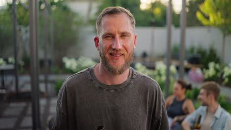 Portrait-of-a-confident-blond-guy-with-a-beard-in-a-gray-T-shirt-holding-a-brown-bottle-of-beer-and-posing-in-the-yard-of-a-back-house-near-his-company-on-vacation