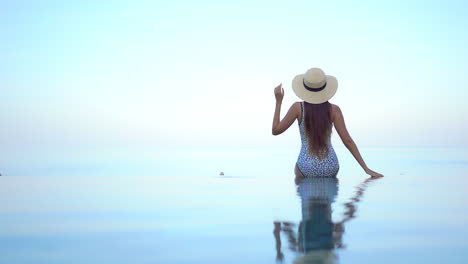 Vista-Trasera-De-Una-Mujer-Con-Traje-De-Baño-Y-Sombrero-Sentada-Al-Borde-De-La-Piscina-Infinita-En-La-Azotea-Con-Reflejo-Corporal-En-El-Agua-Y-El-Cielo-De-La-Puesta-De-Sol,-Plantilla-De-Vacaciones