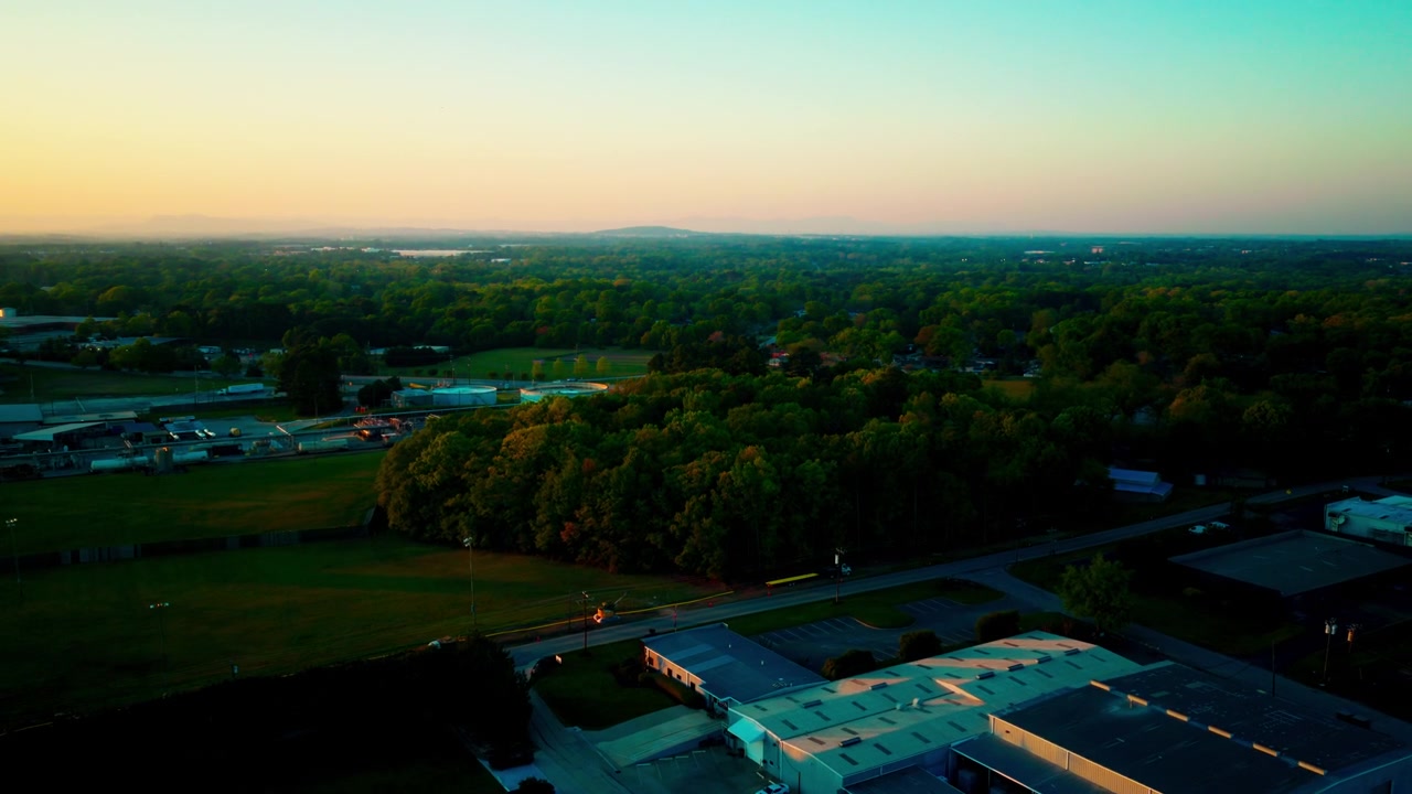 Droneoptagelse i skumringen over Mauldin, South Carolina, der viser et distributionslager med læsserampe, et stort trækronetag og en fjern bjergsilhuet på horisonten i det nordlige Piedmont i den blå time.