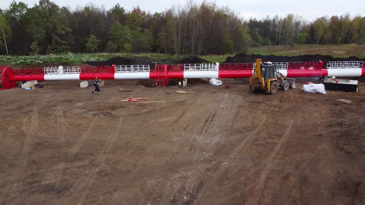 Ampia vista di un cantiere rurale con una lunga sezione di conduttura rossa e bianca che si estende attraverso il sito, un escavatore giallo nelle vicinanze e un operaio presente.