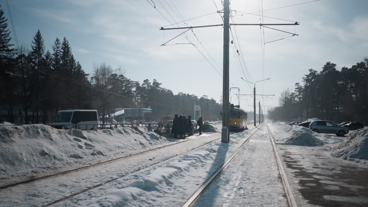 long shot winter street tram approaches stop under catenary, group of people waiting beside snowy platform, cars parked along road, as doors open for boarding on quiet cold morning