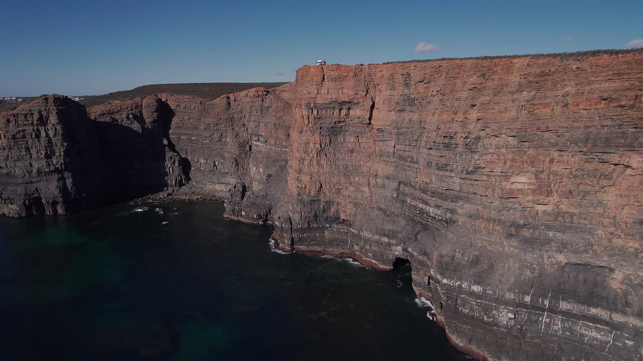 Ampia parete rocciosa sedimentaria orizzontale lungo la costa atlantica a Vila Nova de Mil Fontes, Odemira, Alentejo, Portogallo, con una fascia di erosione visibile e un recesso di grotta marina, ripresa con drone stabile in avvicinamento e ascesa.