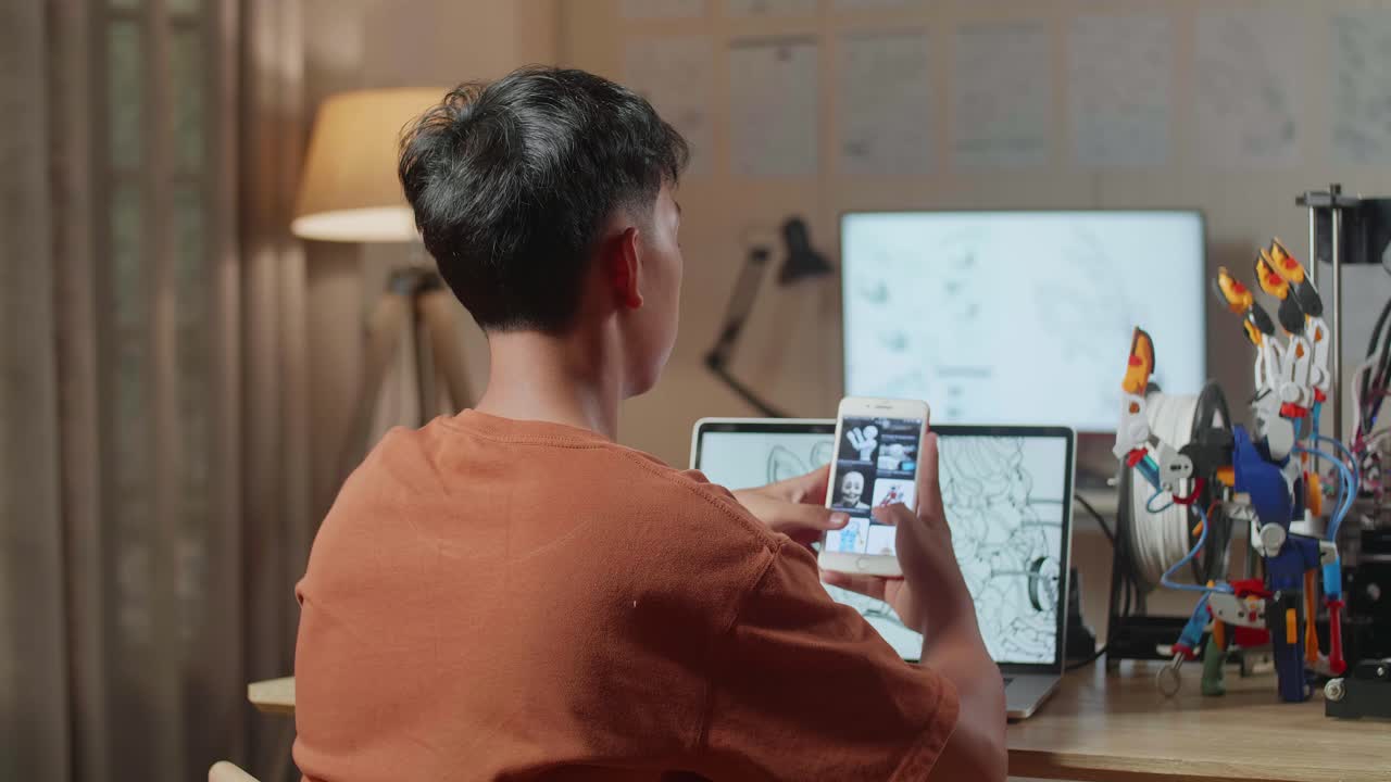 vista trasera de un niño con impresión 3d mirando las imágenes en el teléfono inteligente mientras diseña una mano cyborg en una computadora portátil en casa