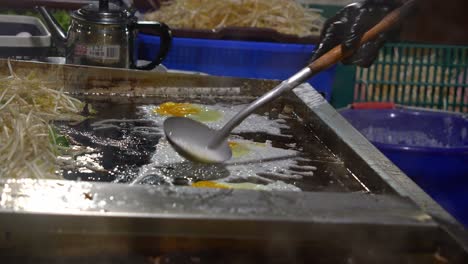 a street food vendor pours a starch mixture and topping it with fresh oysters on the flat-top griddle, cooking a traditional oyster omelette at the bustling local night market, close up shot.