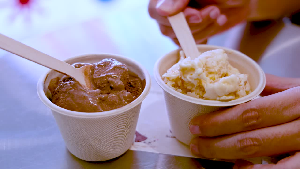 A hand uses a wooden spoon to scoop vanilla ice cream from a paper cup beside a chocolate ice cream cup on a reflective table under soft indoor lighting