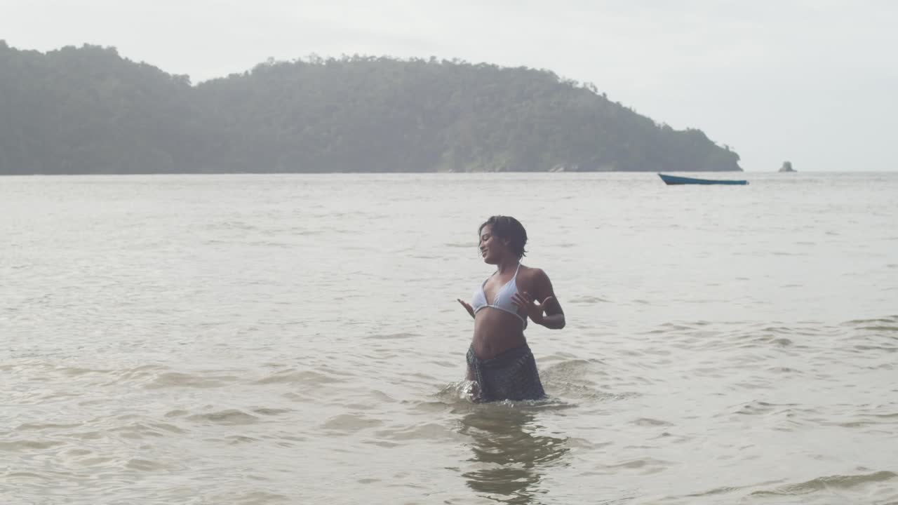 una niña bailando en las aguas tropicales de la isla caribeña de trinidad con montañas y un barco de pesca en el fondo