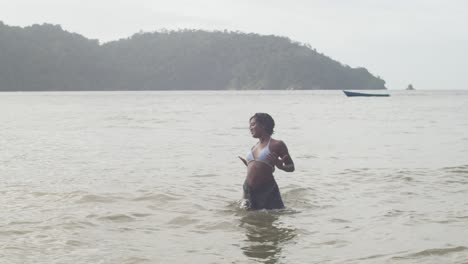 Ein-Junges-Mädchen-Tanzt-Im-Tropischen-Wasser-Der-Karibischen-Insel-Trinidad-Mit-Bergen-Und-Einem-Fischerboot-Im-Hintergrund