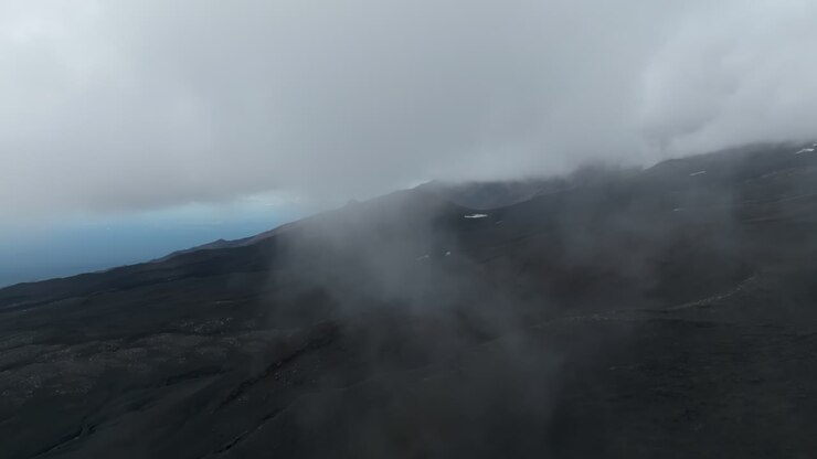 Aerial footage of Tolbachinsky Dol volcanic plateau with dark lava fields and drifting low clouds in Klyuchevskoy Nature Park, Kamchatka Krai, Russia