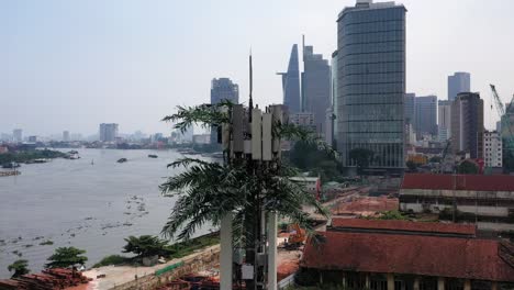 slow panning r to l aerial shot of ultra modern mobile phone tower disguised as a palm tree against a background of a riverfront construction site with city buildings, cranes and boat traffic
