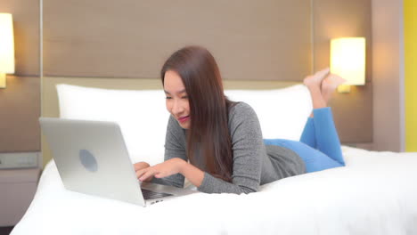 beautiful asian woman lying on the bed with a laptop computer in front of her and typing message on the keyboard smiling