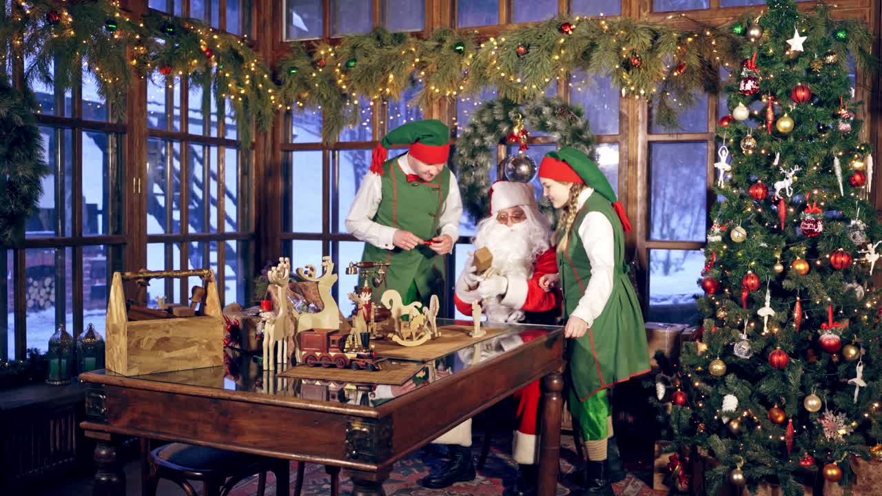 Santa and two elves are getting ready for Christmas in specially decorated room. Girl elf is brinning a plane for wooden presents. New Year and Christmas concept. Feast is coming.