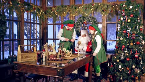 santa and two elves are getting ready for christmas in specially decorated room. girl elf is brinning a plane for wooden presents. new year and christmas concept. feast is coming.