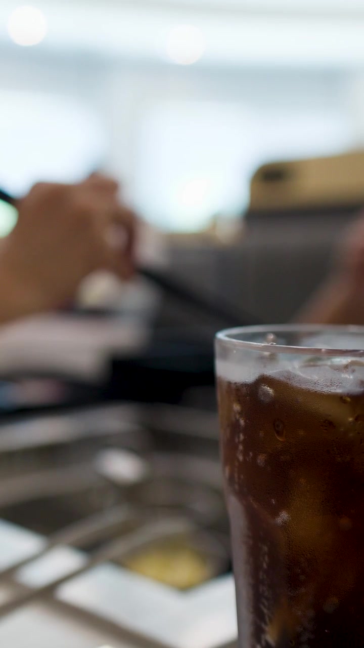 Un bicchiere alto di cola ghiacciata è su un tavolo mentre i commensali mangiano sullo sfondo.