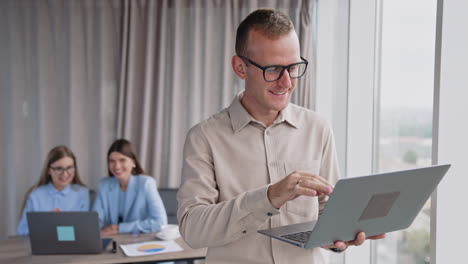 man wearing glasses holds a laptop standing near window and has video call. a pair of female colleagues sit at desk and laugh looking at computer at backdrop in blur.