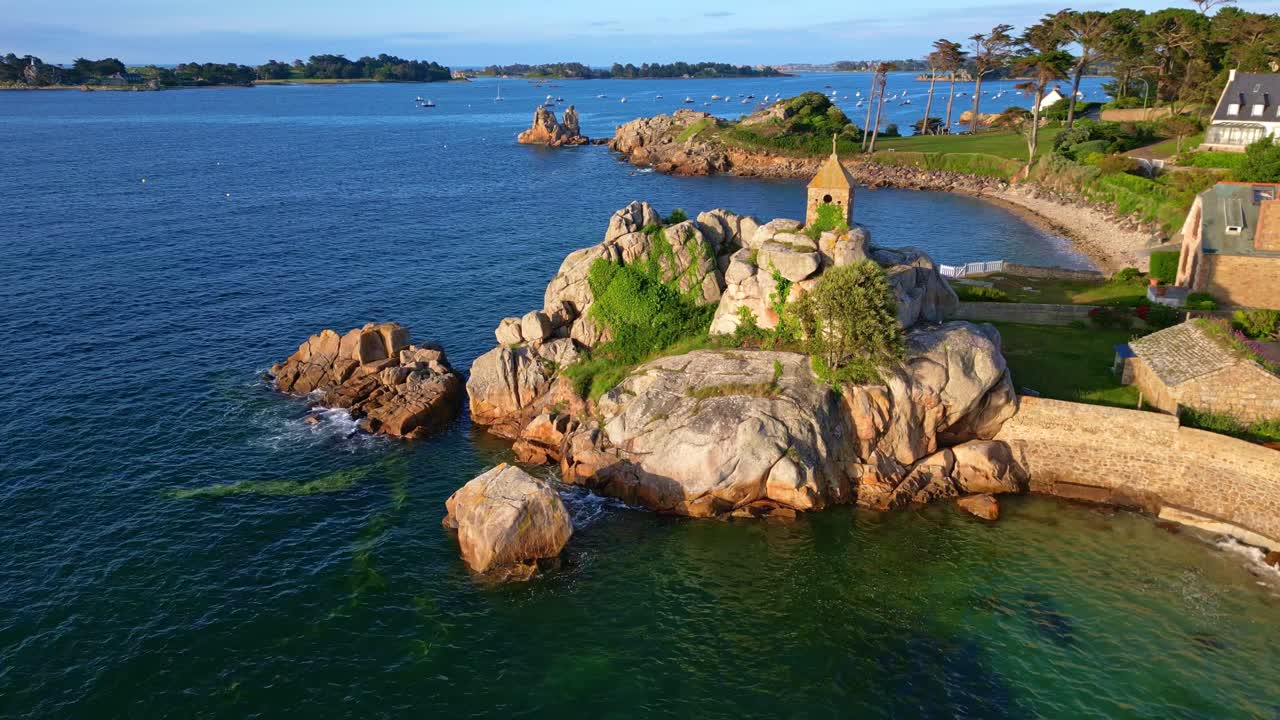 Drone moves back from Sentinelle oratory, revealing nearby houses, rocky shore, sea, and Port-Blanc bay.