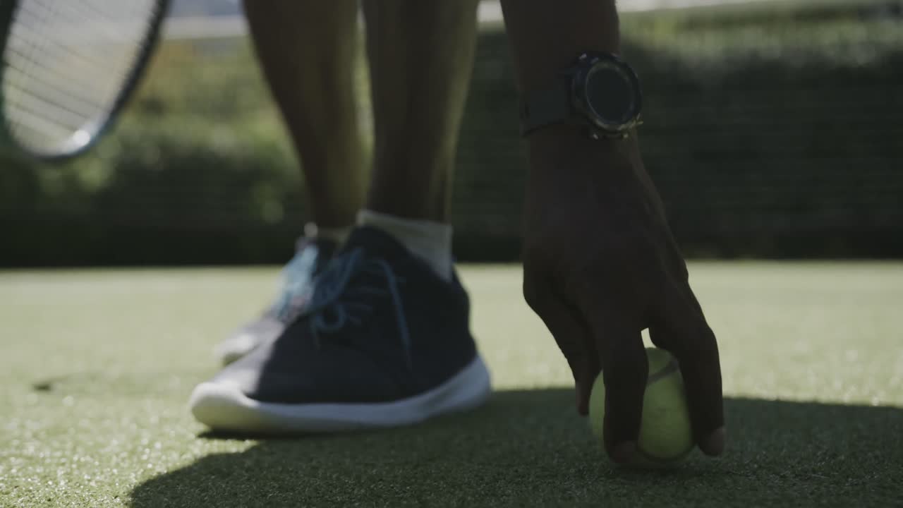 sección baja de una mujer afroamericana senior recogiendo una pelota de tenis en la cancha de tenis en cámara lenta