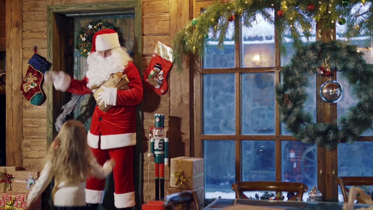 Santa Claus opens the door and comes inside his residence. Happy children and fairy elves running to Santa in Christmas decorated room.