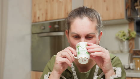 woman in green dress enjoying peaceful breakfast moment while sipping hot beverage from vintage ceramic cup in bright kitchen surrounded by warm wood accents and soft natural lighting