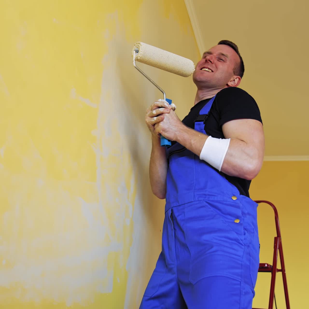 Funny craftsman painting walls. Cheerful young male worker imitating singing on a paint roller during his work on room renovation