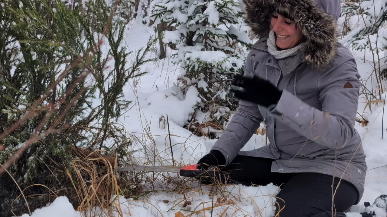 feliz, mujer sonriente cortando un árbol de navidad en un bosque nevado con una sierra