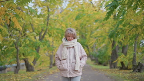 middle age woman in soft pink jacket and thick knitted scarf stands calmly with hands tucked in pockets under vibrant yellow and green trees along quiet park path scattered with fallen leaves