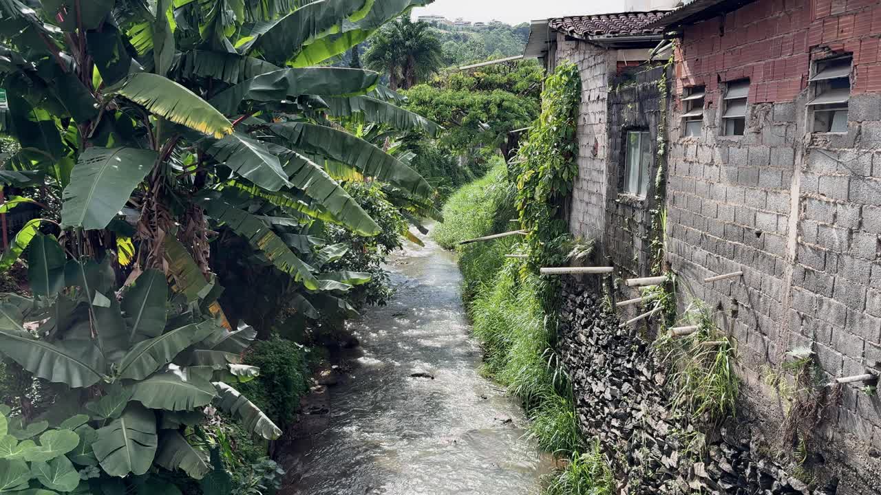 4K image of a canal in Viçosa, captured from a bridge. The scene includes lush greenery surrounding the waterway and modest houses on the side, blending nature with urban simplicity.
