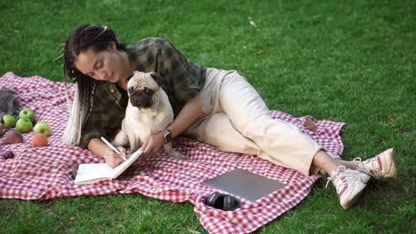 chica elegante acostada en cuadros en el césped en un parque y haciendo notas mientras un pequeño pug sentado a su lado, portátil, auriculares