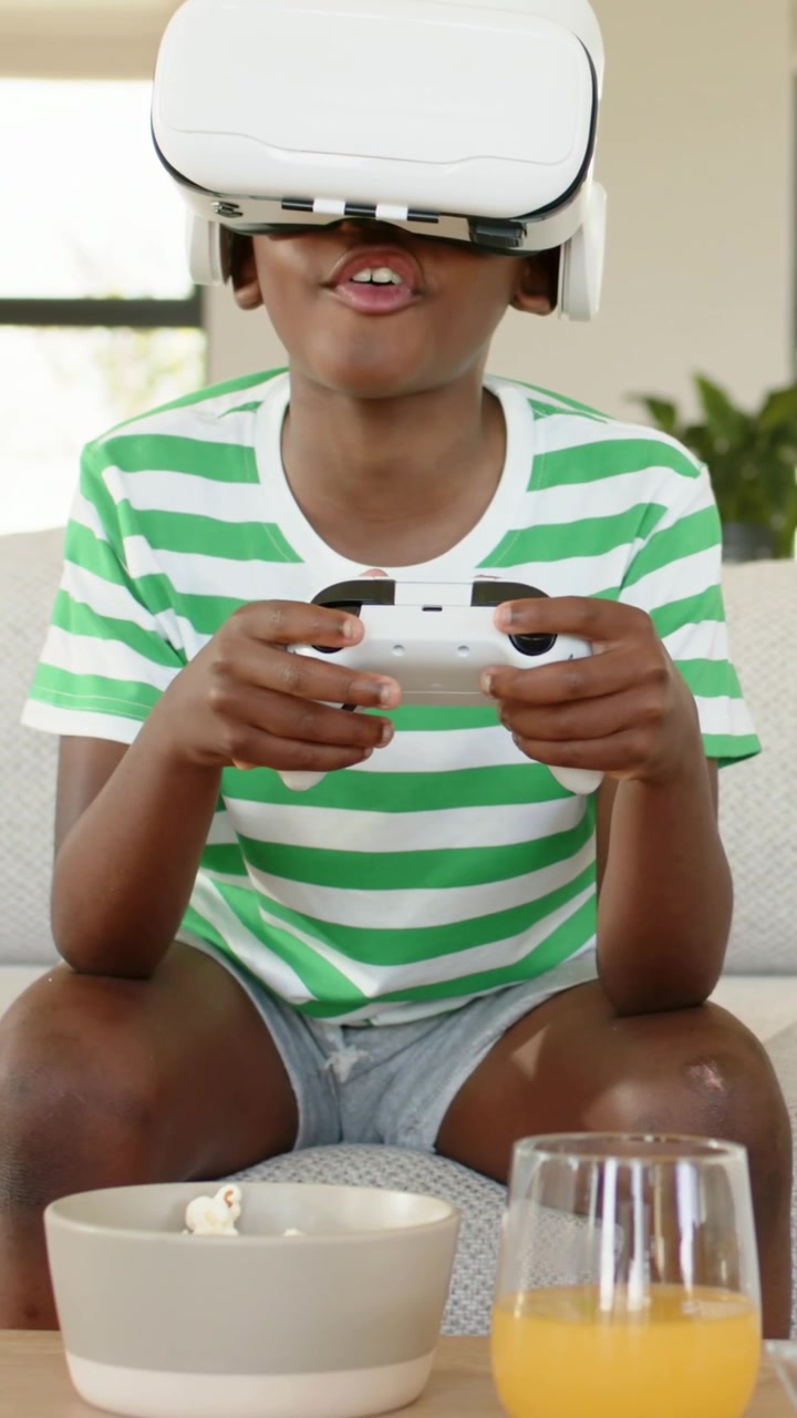 Vertical video: Playing boy in shirt pressing controller on sofa as headset showing, popcorn, juice