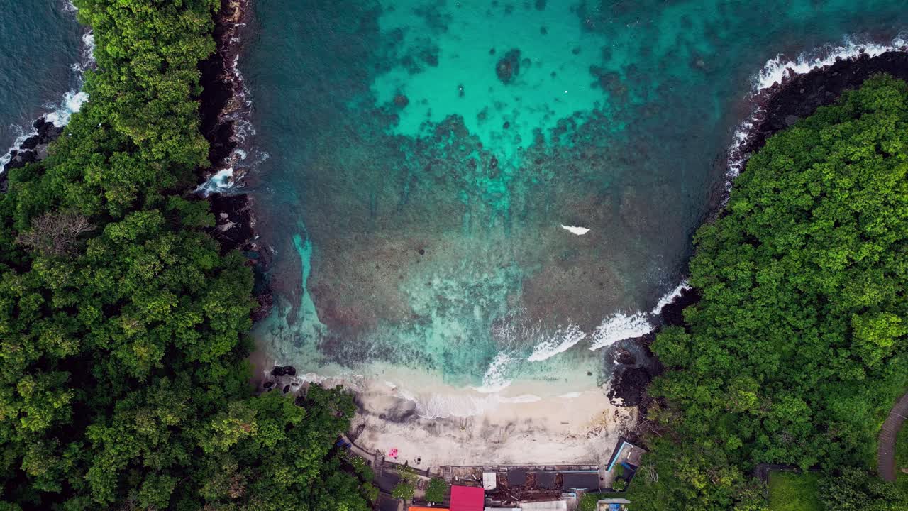 Overhead drone shot of Blue Lagoon Beach in East Bali, revealing vibrant turquoise waters, coral reefs, and a secluded white sand bay surrounded by thick tropical vegetation and cliffs