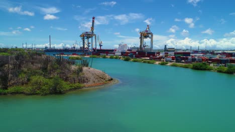 Aerial-flyover-of-shipping-container-port-with-cranes-and-boat-docked,-sunny-day-on-Curacao-Caribbean