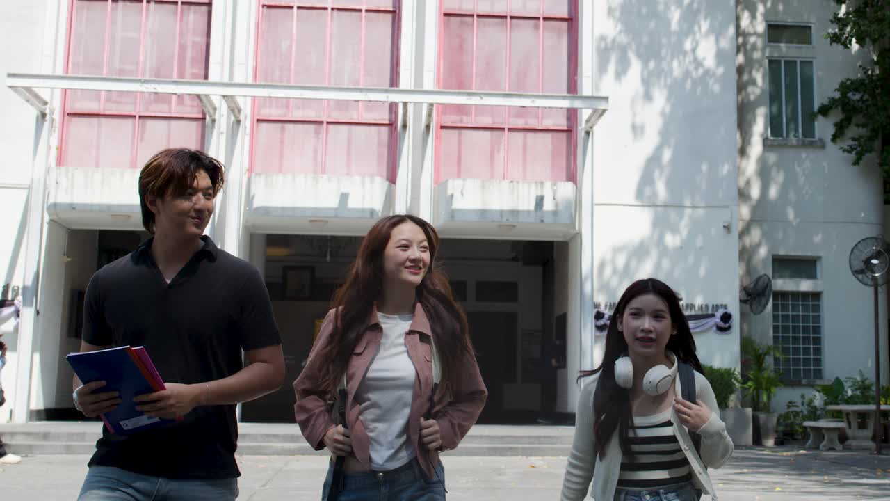 Tre studenti asiatici camminano insieme fuori da un edificio universitario, portando libri e parlando allegramente.