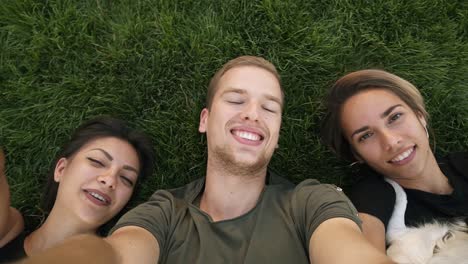 three attractive millennial friends take selfies while lying on the green grass in the park or lawn