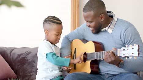 Glücklicher-Afroamerikanischer-Vater-Und-Sohn-Sitzen-Auf-Dem-Sofa-Und-Spielen-Gitarre,-In-Zeitlupe