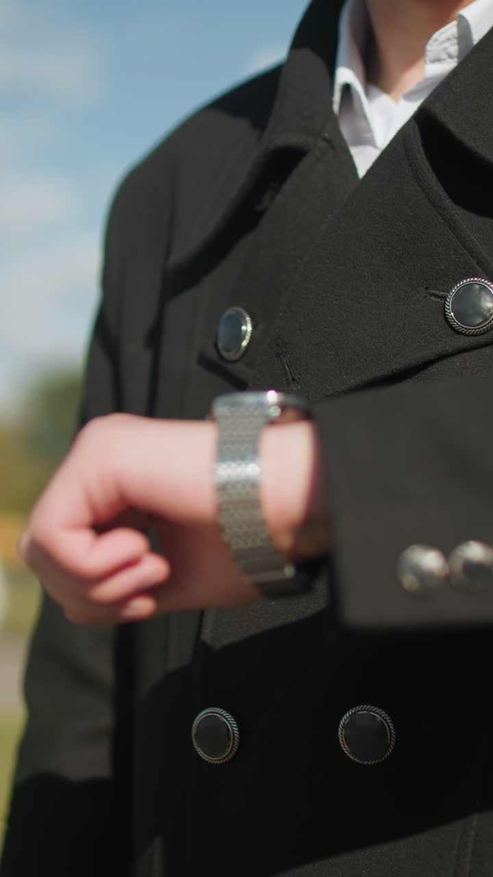 primer plano de un inversor caminando con confianza al aire libre comprobando la hora en un reloj de pulsera, con un elegante abrigo negro con botones metálicos, en contra de la arquitectura urbana moderna, árboles verdes y un cielo azul claro