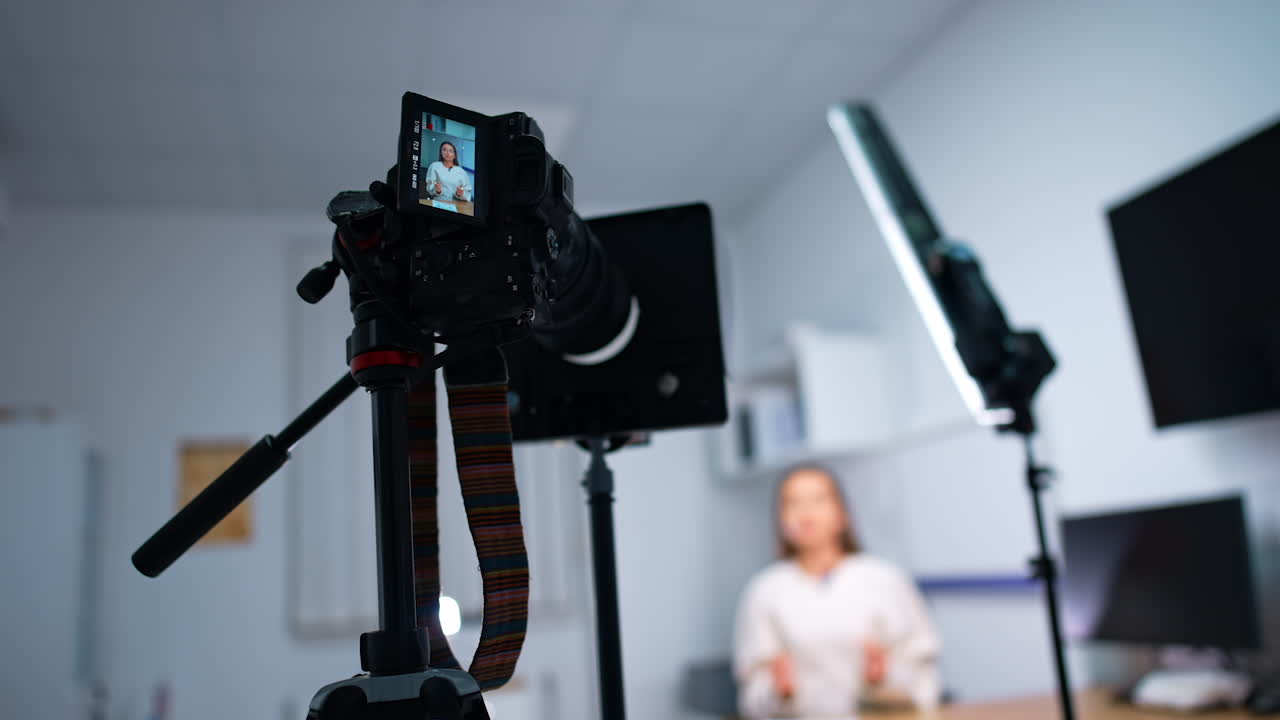 Camera set on tripod recording a female blogger. Long-haired woman talks to camera. Low angle view. Blurred backdrop.