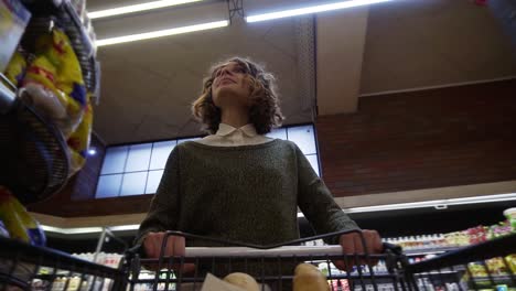 pretty woman in casual clothes is walking in grocery store steering shopping trolley with food inside it and looking around at shelves with products. women and shops concept. low angle footage from the cart