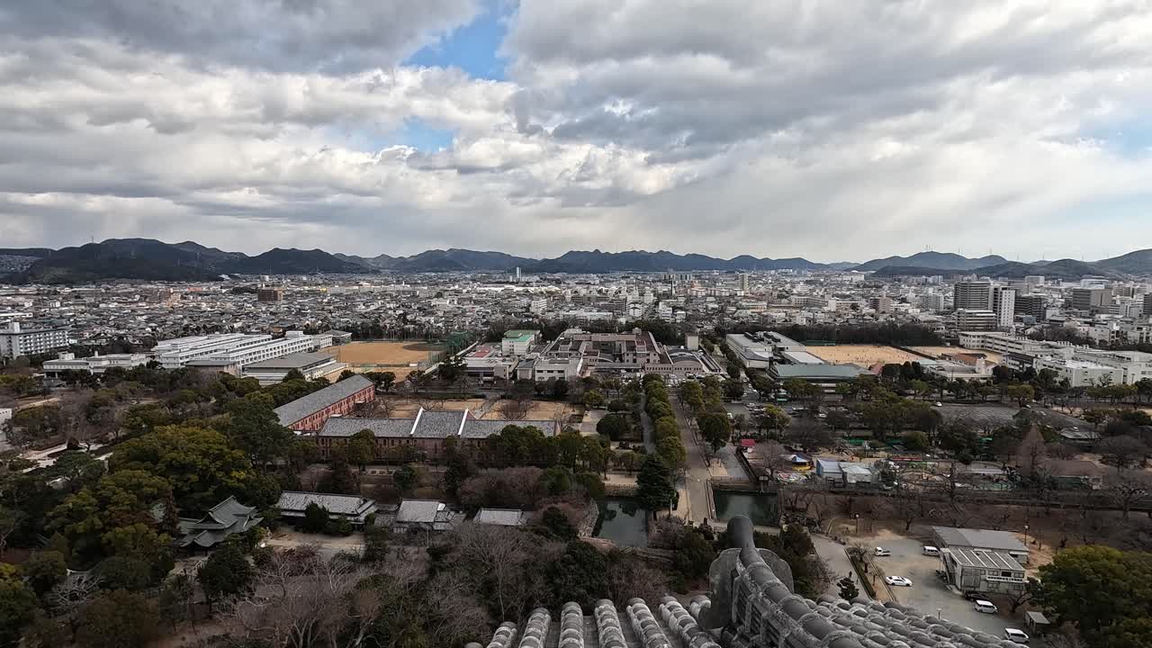 Captured from the top of Himeji Castle, this panoramic view showcases the blend of traditional tiled rooftops, tree-lined avenues, and the expansive city of Himeji under a dramatic sky.